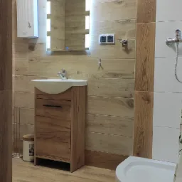 Nowoczesna łazienka w Siemiatyczach z drewnopodobnymi płytkami, podświetlanym lustrem i kabiną prysznicową z białym brodzikiem.