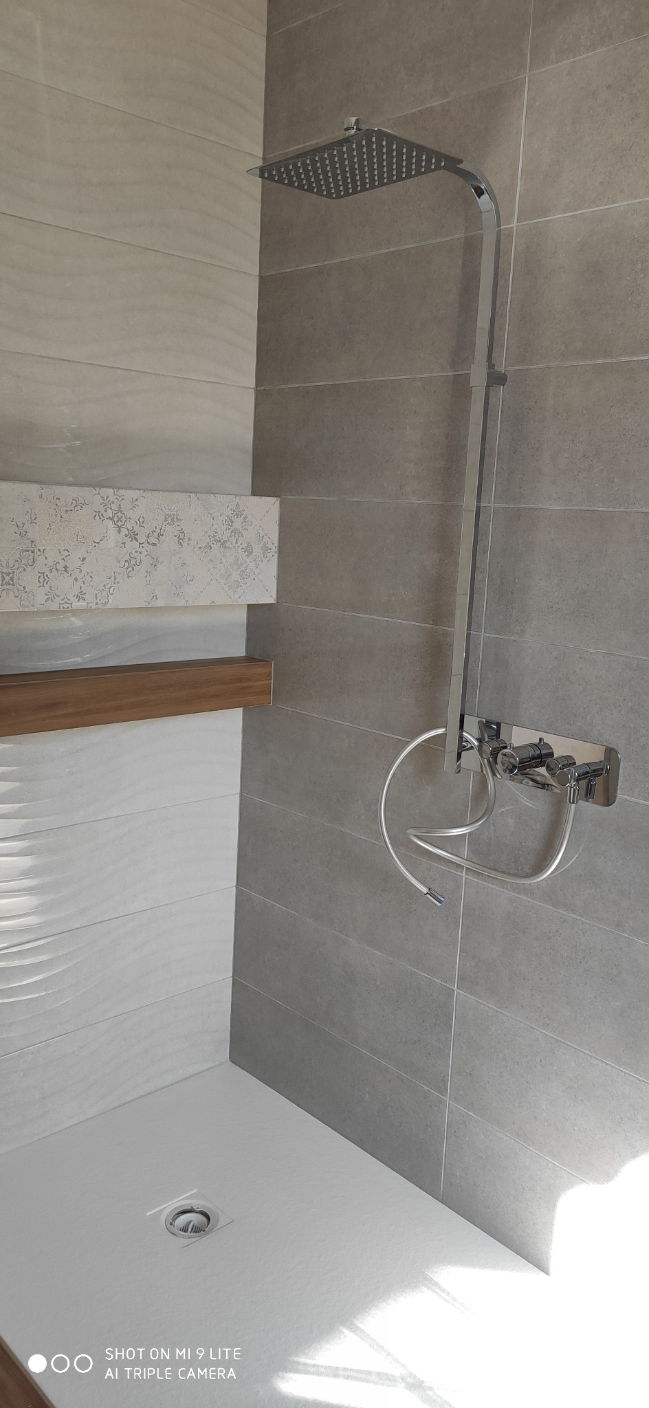 Nowoczesna kabina prysznicowa z chromowaną armaturą, szarymi płytkami i dekoracyjnym panelem ściennym z drewnianą półką.
