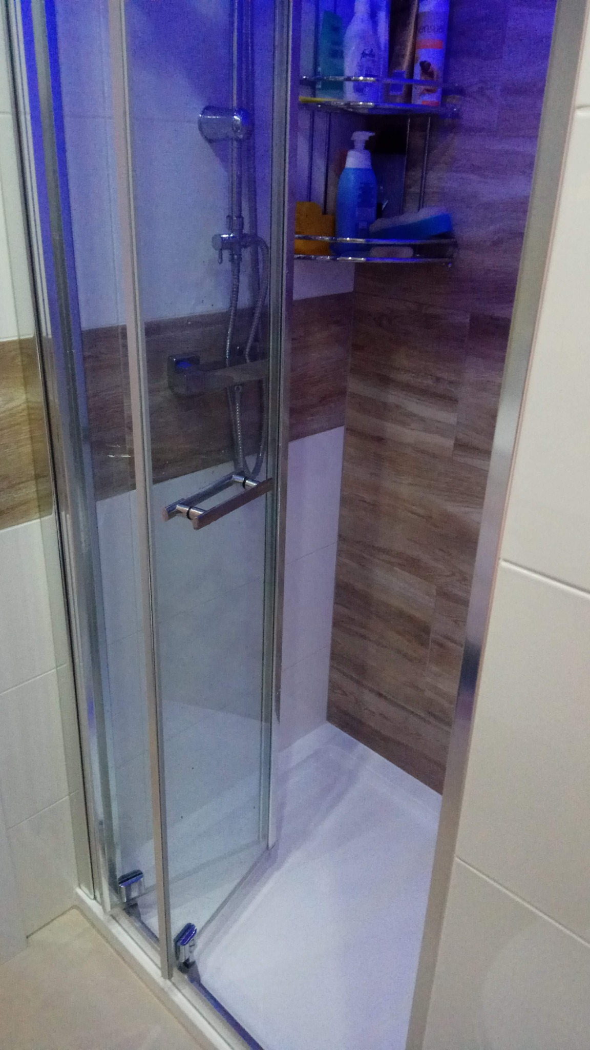 Szklana kabina prysznicowa z chromowanymi elementami, widoczna półka z kosmetykami i brązowe płytki imitujące drewno na ścianie, oświetlona niebieskim światłem.