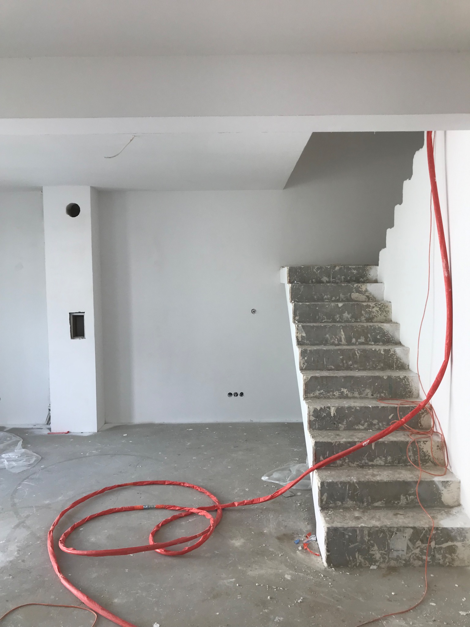 Surowe betonowe schody w trakcie budowy w jasnym pomieszczeniu, z widoczną czerwoną rurą grzewczą i surowymi ścianami przygotowanymi do malowania.