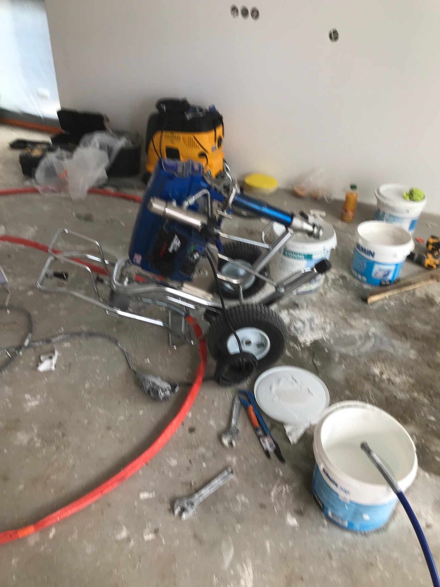 Agregat malarski z wężami i wiadrami farby na zanieczyszczonej podłodze w trakcie prac wykończeniowych wnętrza.