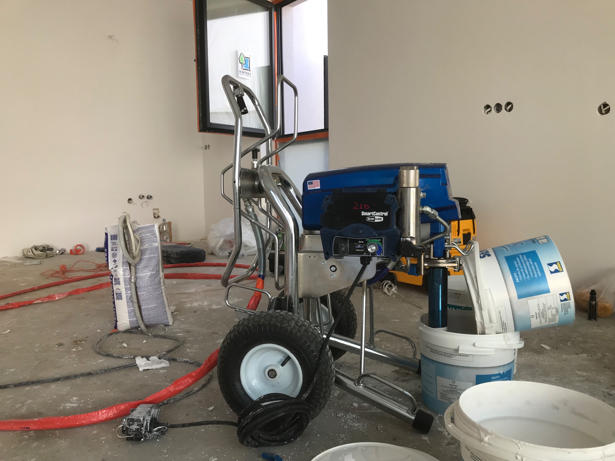 Agregat malarski z wężem i wiadrami farby na podłodze w trakcie prac wykończeniowych, widoczne puszki farby marki Semin oraz niedomalowane ściany z otworami na gniazdka elektryczne.