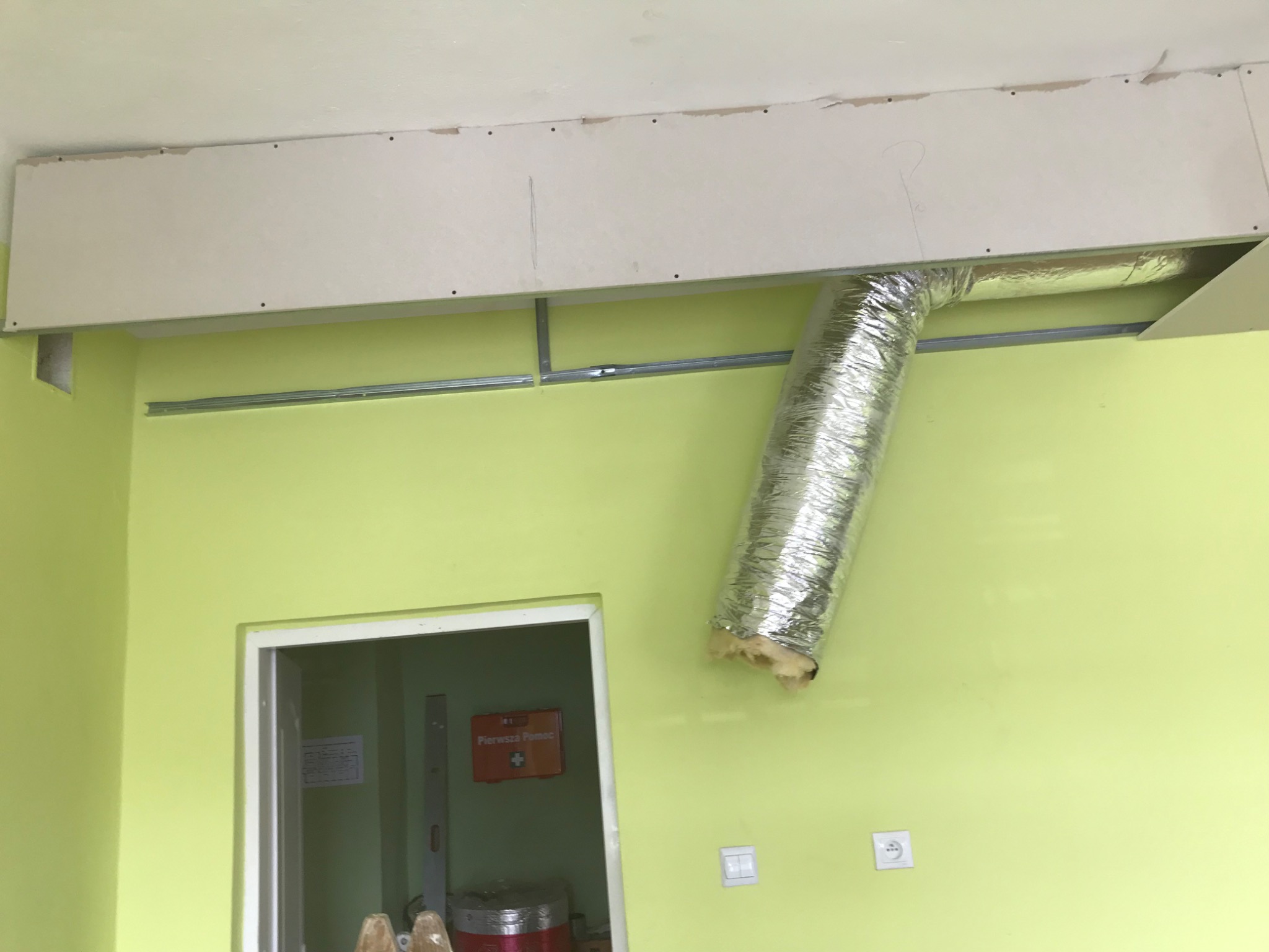 Montaż instalacji wentylacyjnej z widoczną rurą aluminiową, częściowo zakrytą płytą kartonowo-gipsową w pomieszczeniu o jasnozielonych ścianach, widoczna apteczka pierwszej pomocy w tle.