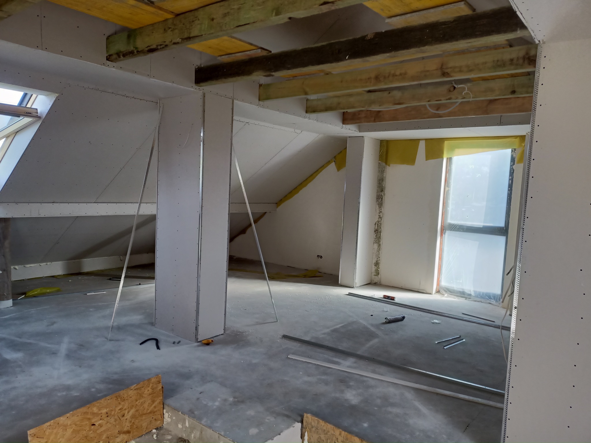 Poddasze w trakcie adaptacji: widoczne drewniane belki stropowe, montowane płyty gipsowo-kartonowe na ścianach i skosach, profile metalowe, okno dachowe. W tle widoczna żółta izolacja.