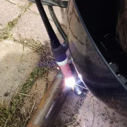 Spawanie rury metalowej metodą TIG w warunkach polowych, widoczny jasny łuk elektryczny oraz maska spawalnicza.