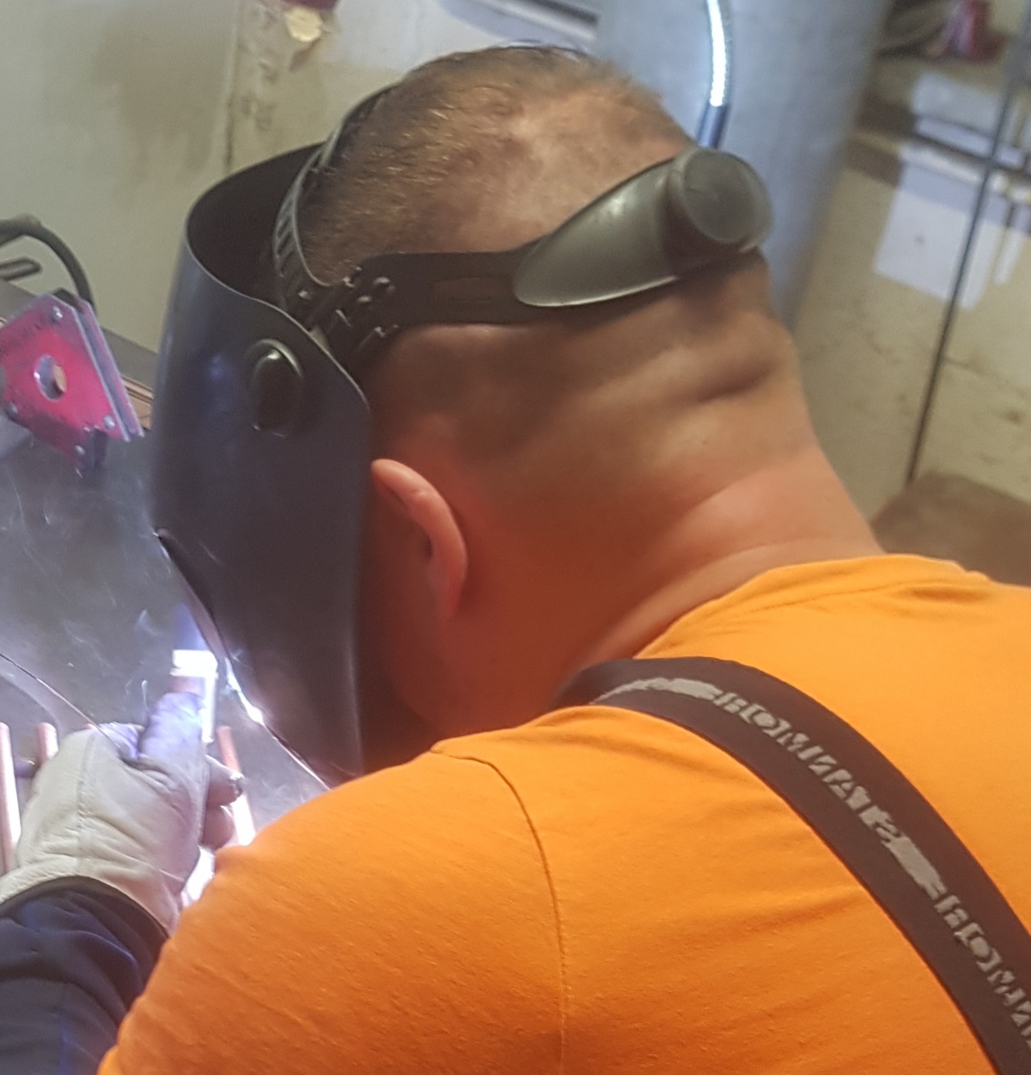 Spawanie metalowych elementów konstrukcyjnych w warsztacie, widok zza pleców spawacza w masce ochronnej i rękawicach, iskry i dym podczas pracy.