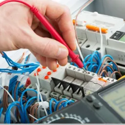 Elektryk mierzy napięcie w rozdzielnicy elektrycznej za pomocą multimetru z czerwoną sondą, widoczne liczne przewody w kolorze niebieskim i brązowym.