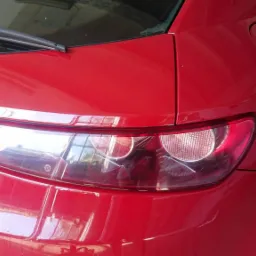 Czerwony samochód typu hatchback z widocznym tylnym światłem i wycieraczką, odbicie światła na lakierze.