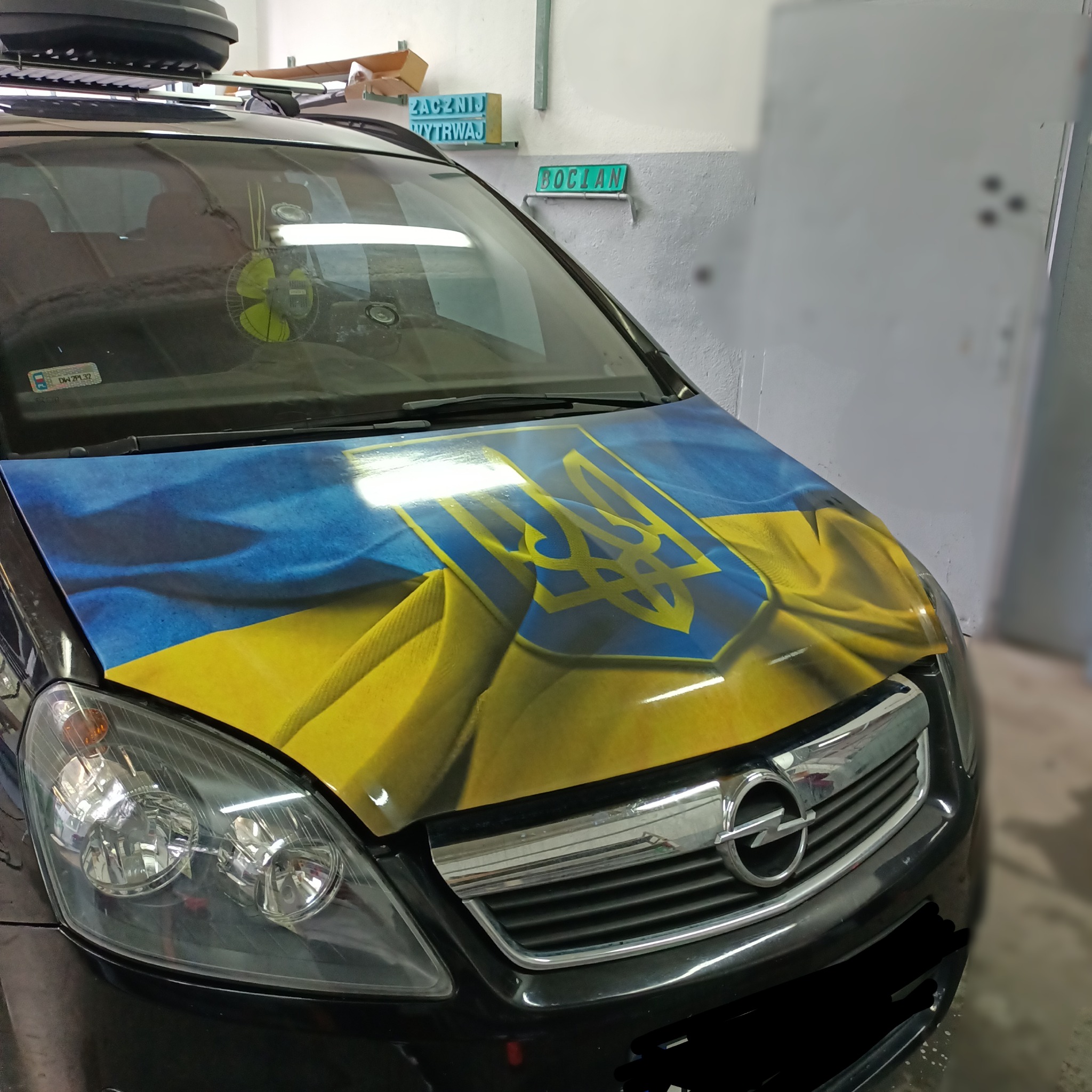 Czarny samochód Opel Zafira z oklejoną maską w barwy flagi Ukrainy i godłem na środku, widoczne reflektory, grill i fragment zderzaka, w tle garaż.