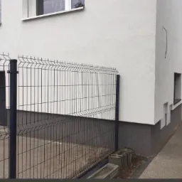 Nowo postawione, czarne, metalowe ogrodzenie panelowe na tle białej elewacji budynku z szarym cokołem i otwartymi, czarnymi drzwiami wejściowymi.