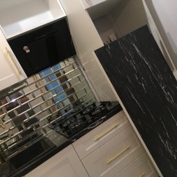 Przemek Snochowski - Nowoczesna kuchnia z lustrzanymi płytkami, czarnym blatem i złotymi uchwytami. Widoczna płyta gazowa i okap. Górne szafki białe, dolne w odcieniach szarości.