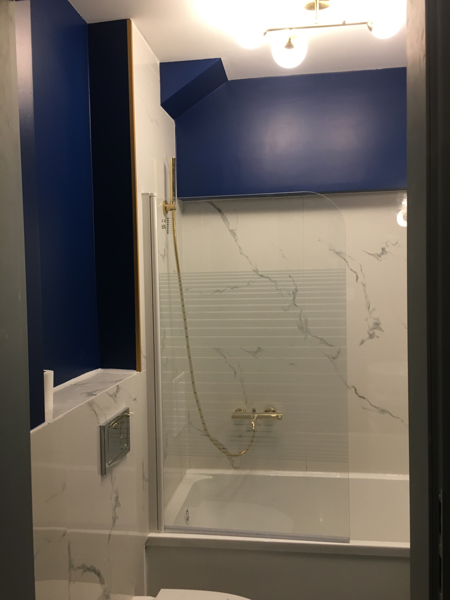 Wykończona łazienka w bieli i granacie: marmurowe płytki, szklany parawan nawannowy z złotą armaturą, oświetlenie sufitowe.