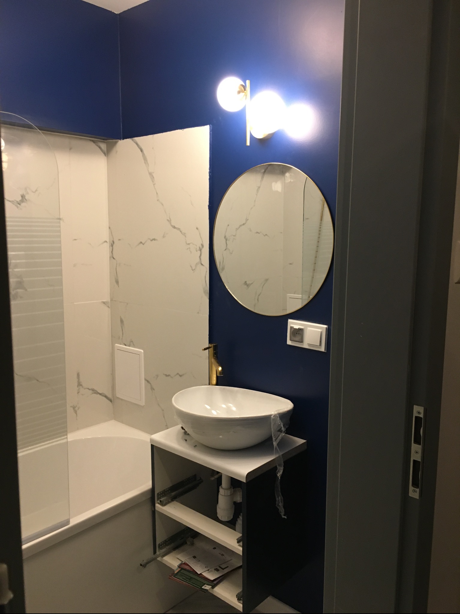 Nowoczesna łazienka po remoncie: biała umywalka na szafce, okrągłe lustro z podświetleniem na granatowej ścianie, białe płytki imitujące marmur w strefie kąpieli.