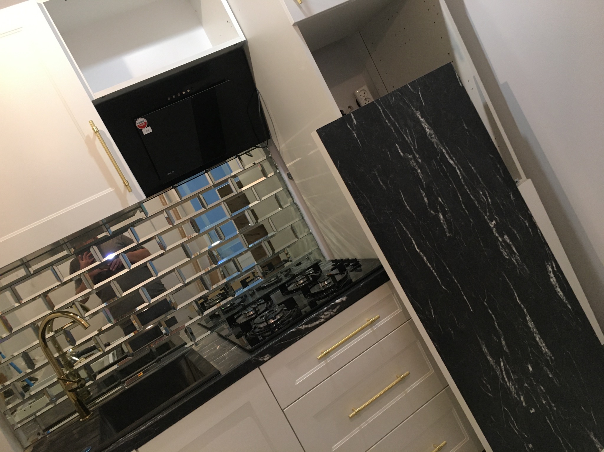 Nowoczesna kuchnia z lustrzanymi płytkami, czarnym blatem i złotymi uchwytami. Widoczna płyta gazowa i okap. Górne szafki białe, dolne w odcieniach szarości.