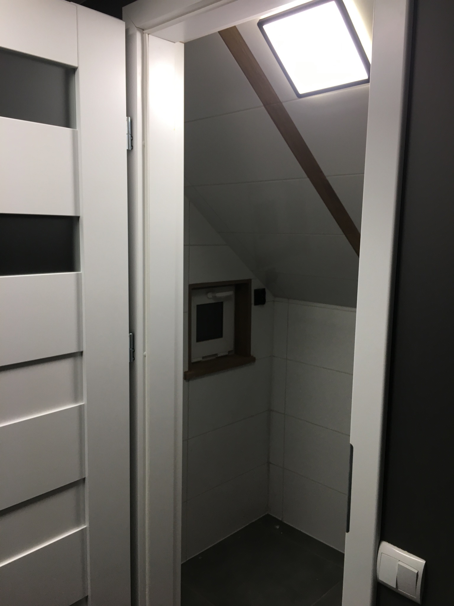 Wykończona łazienka na poddaszu z białą glazurą i oknem. Widok zza białych drzwi z dekoracyjnymi frezami. Oświetlenie sufitowe. Ciemna ściana po prawej.