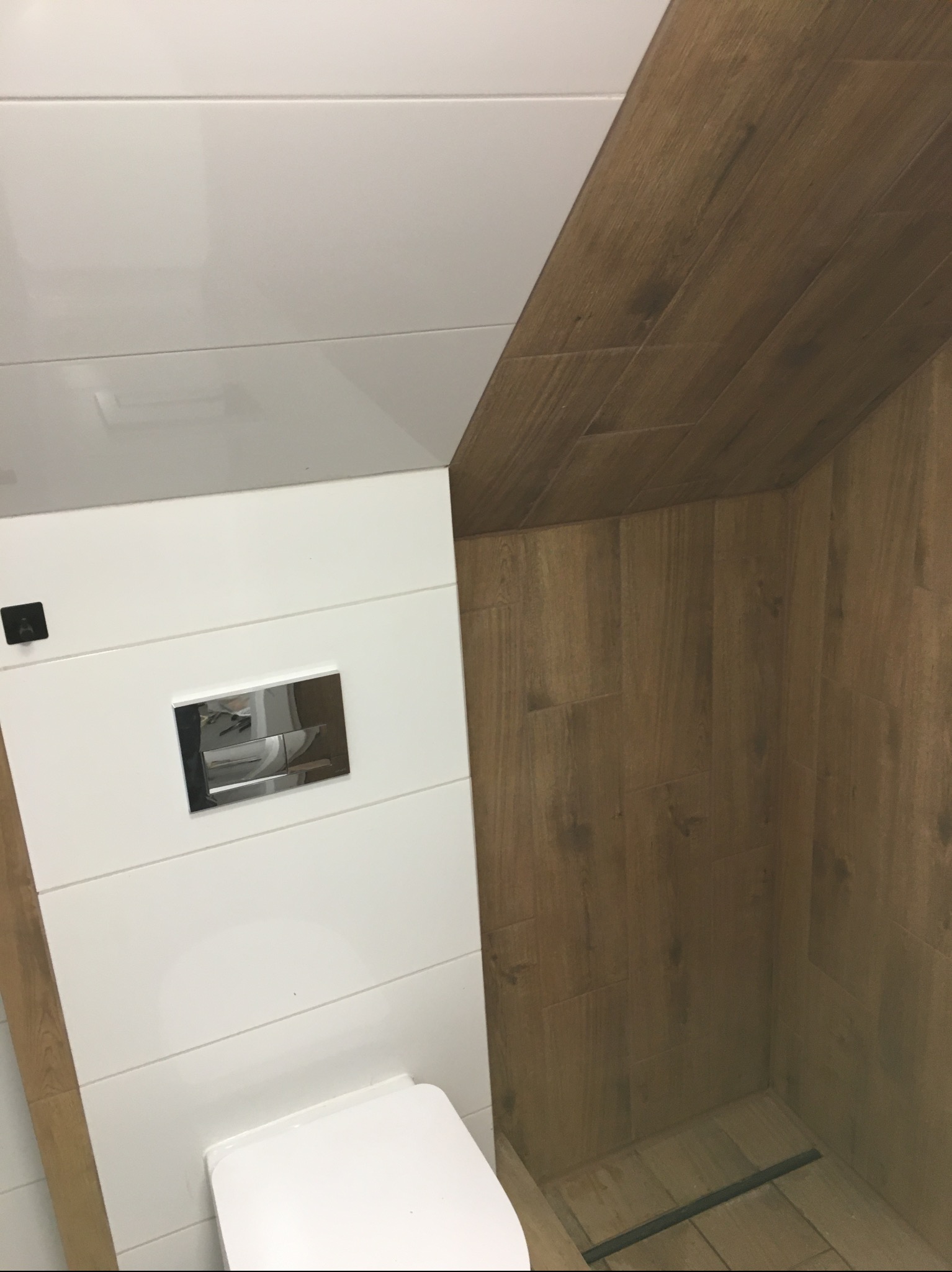 Wykończona łazienka ze stelażem WC obudowanym białymi płytkami imitującymi panele oraz kabiną prysznicową wyłożoną płytkami drewnopodobnymi, z odpływem liniowym.