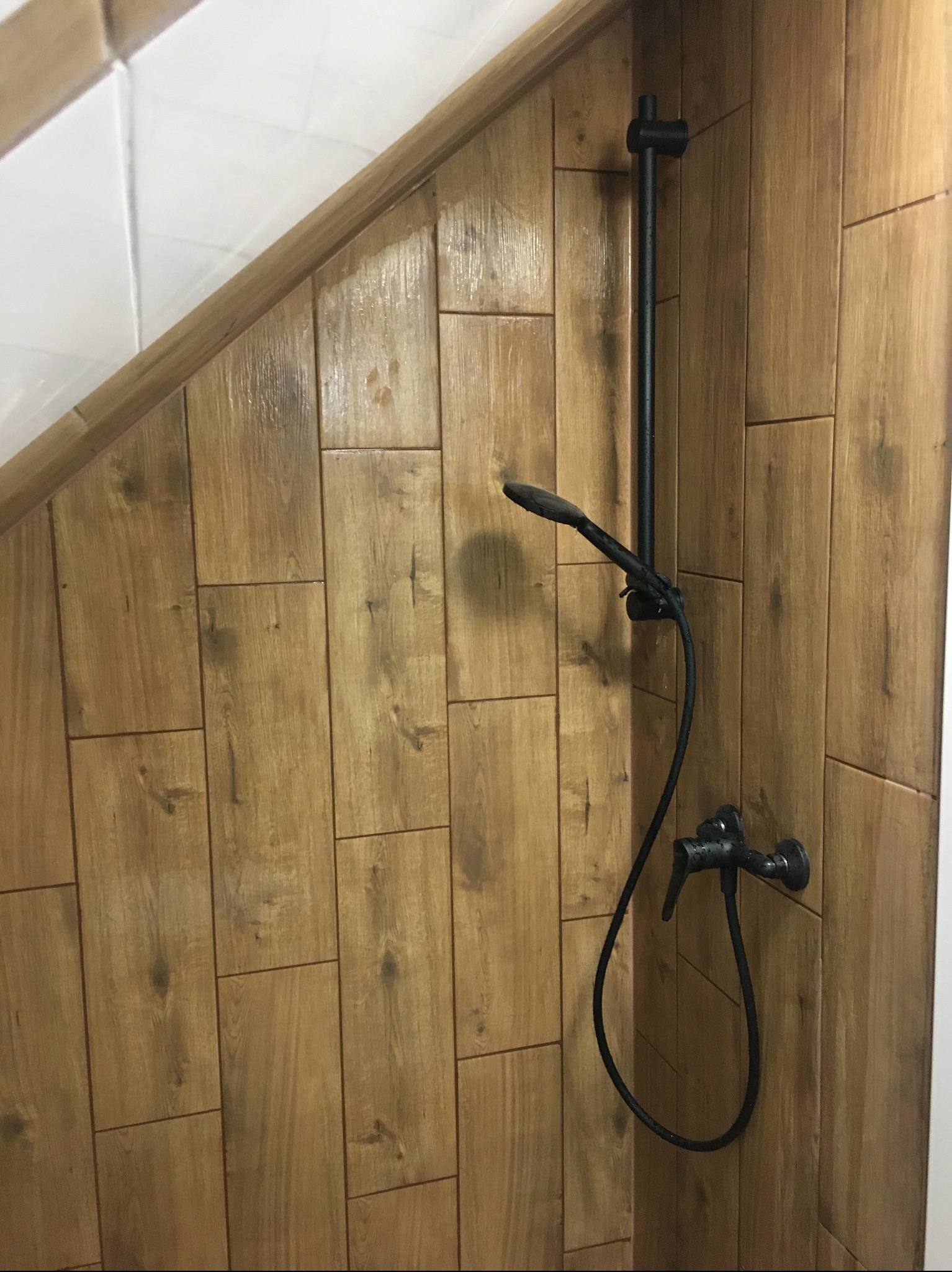 Kabina prysznicowa wyłożona płytkami imitującymi drewno, z czarną armaturą. Nietypowe skosy i połączenie z białymi płytkami dodają unikalnego charakteru.