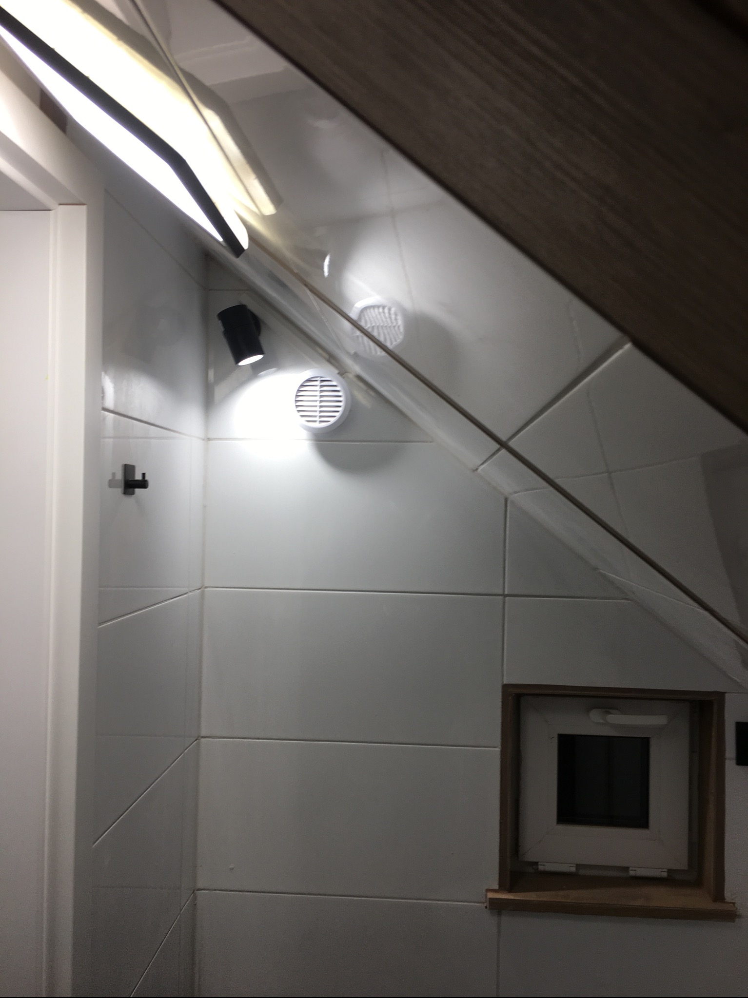 Wykończona łazienka na poddaszu w Koluszkach: białe płytki, okno w drewnianej ramie, oświetlenie punktowe i wentylacja. Ukośny sufit dodaje unikalnego charakteru.