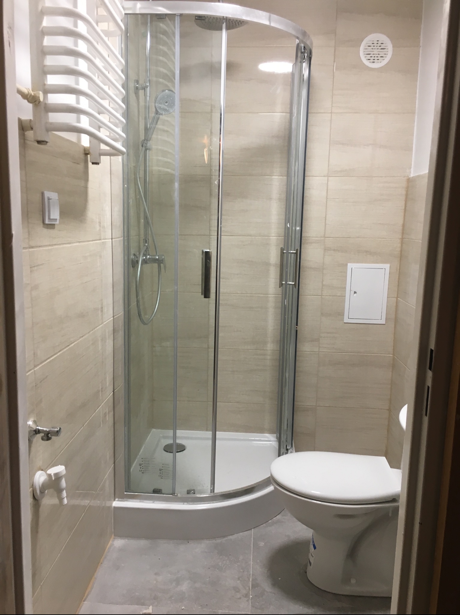 Jasna łazienka po remoncie: kabina prysznicowa z hydromasażem, grzejnik drabinkowy i toaleta. Beżowe płytki na ścianach, szare płytki na podłodze.