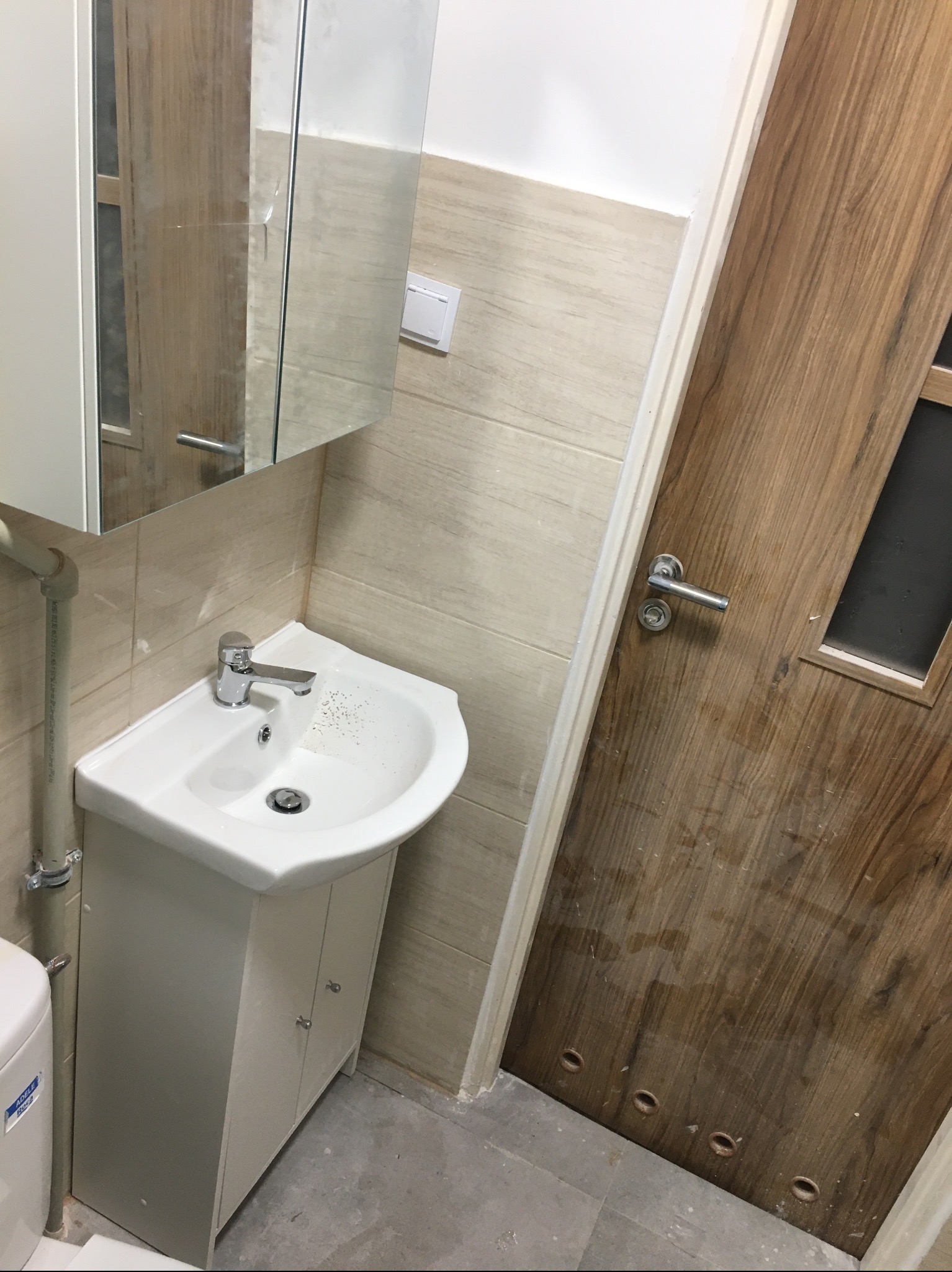 Umywalka z szafką w jasnej łazience z beżowymi płytkami i drewnianymi drzwiami z szybką. Bateria umywalkowa chromowana, lustro nad umywalką.