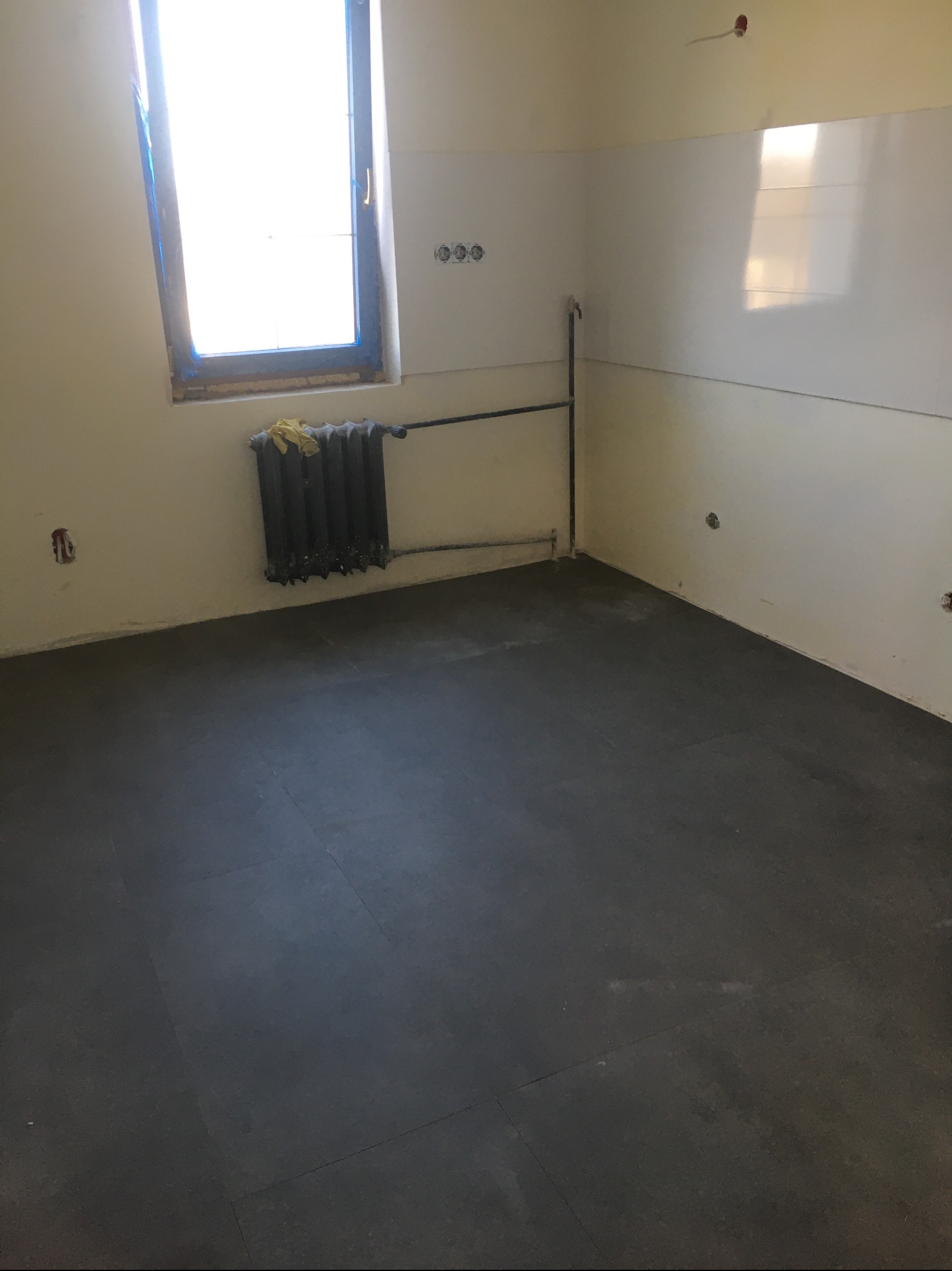Ciemna podłoga z płytek w jasnym pomieszczeniu w trakcie prac wykończeniowych. Widoczny grzejnik pod oknem i przygotowane ściany pod malowanie.