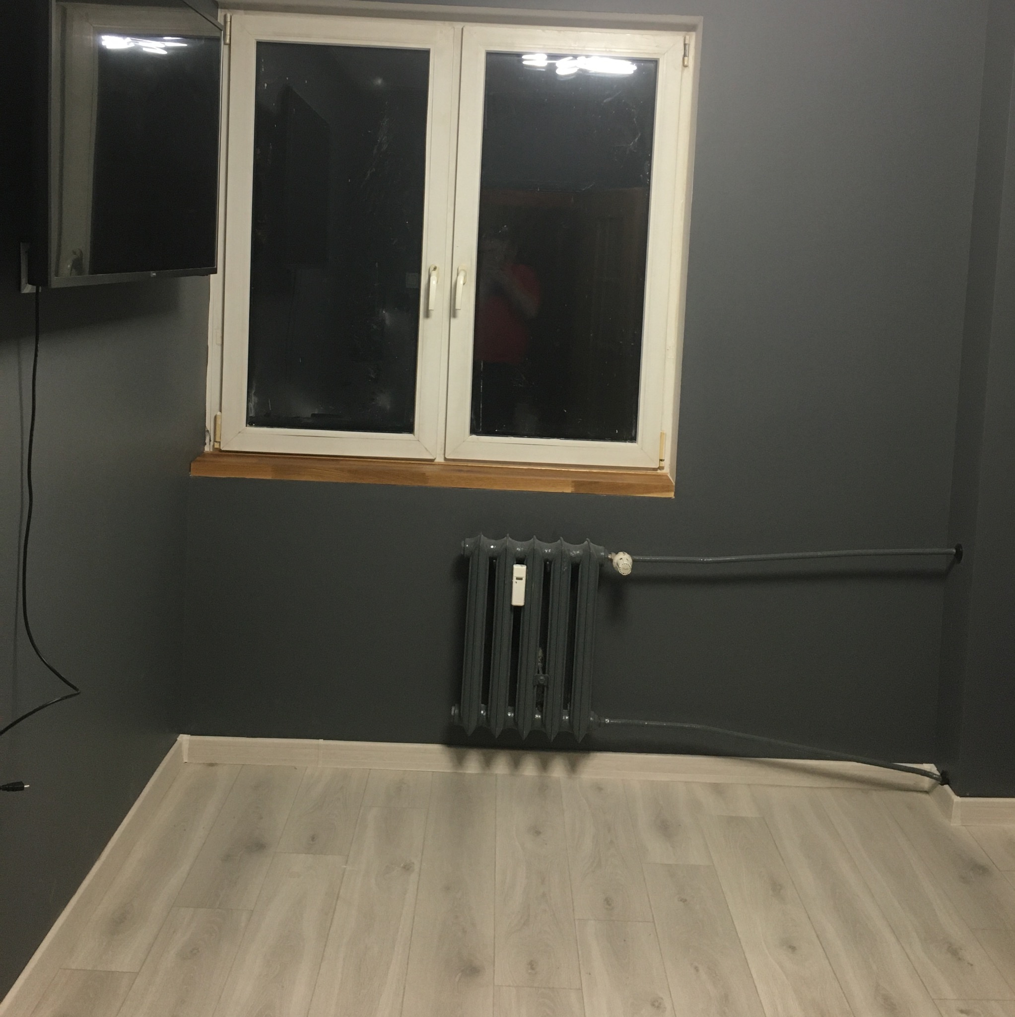 Szara ściana z białym oknem i grzejnikiem, jasne panele podłogowe. Widoczny telewizor na ścianie. Minimalistyczne wnętrze po malowaniu.