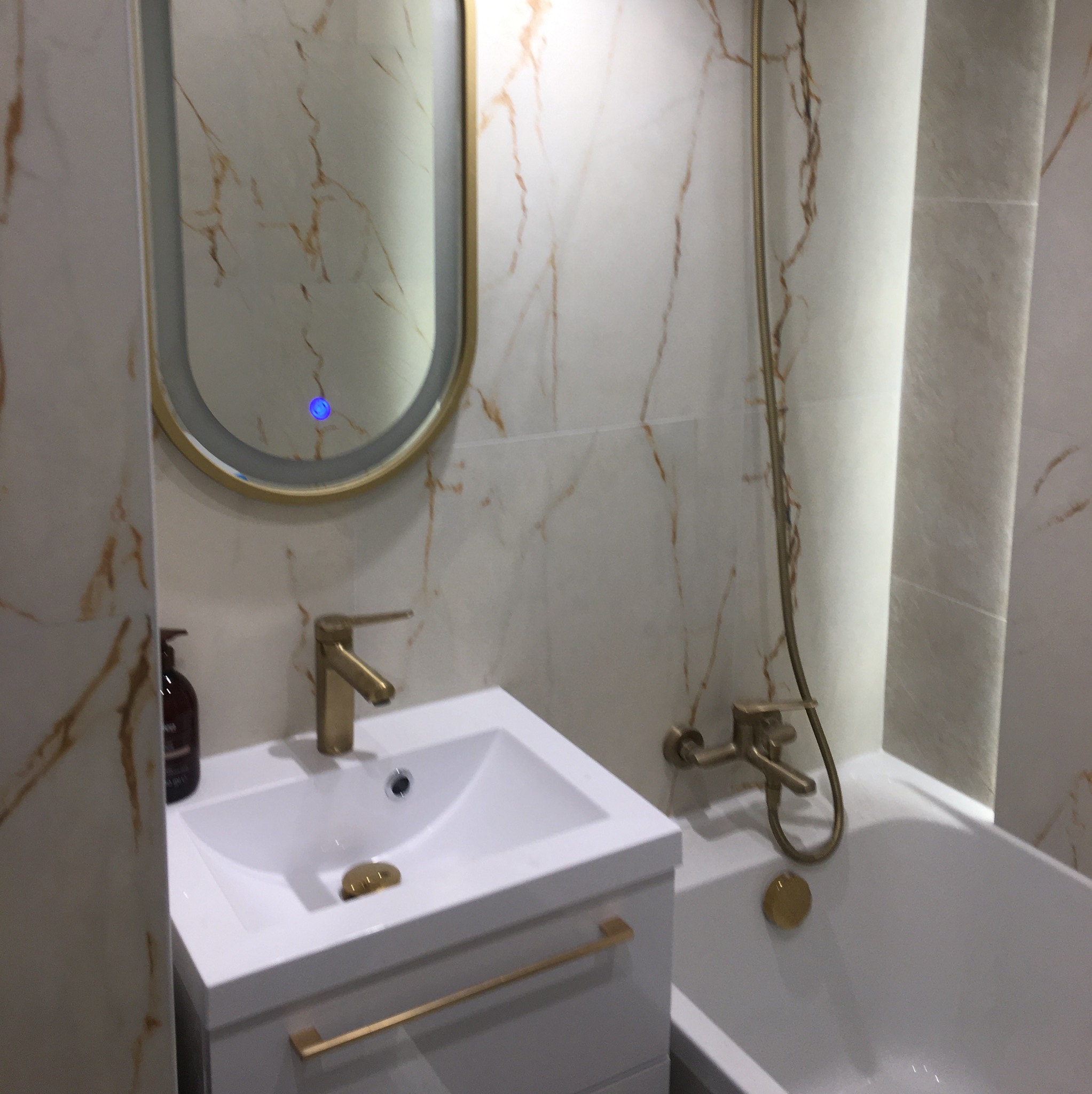 Wykończona łazienka z wanną, umywalką i owalnym lustrem z podświetleniem LED. Armatura w kolorze złotym, płytki imitujące marmur z brązowymi żyłami.