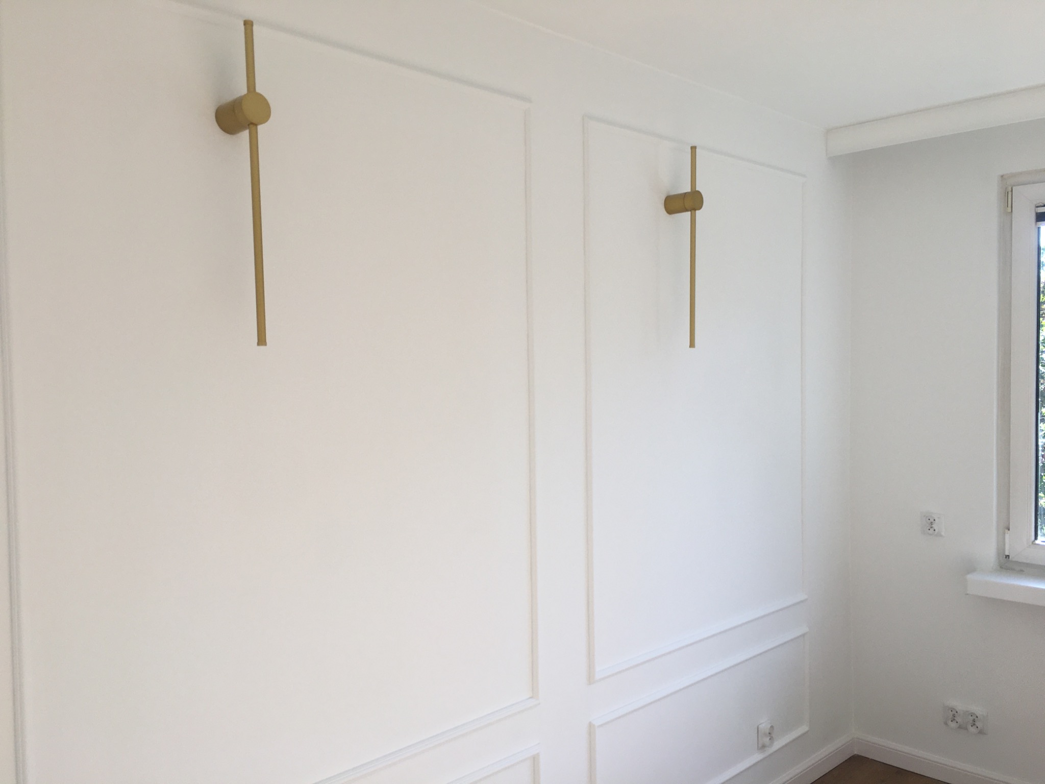 Biała ściana z dekoracyjnymi panelami i dwoma złotymi lampami ściennymi, widok na fragment okna z prawej strony. Minimalistyczny design wnętrza.