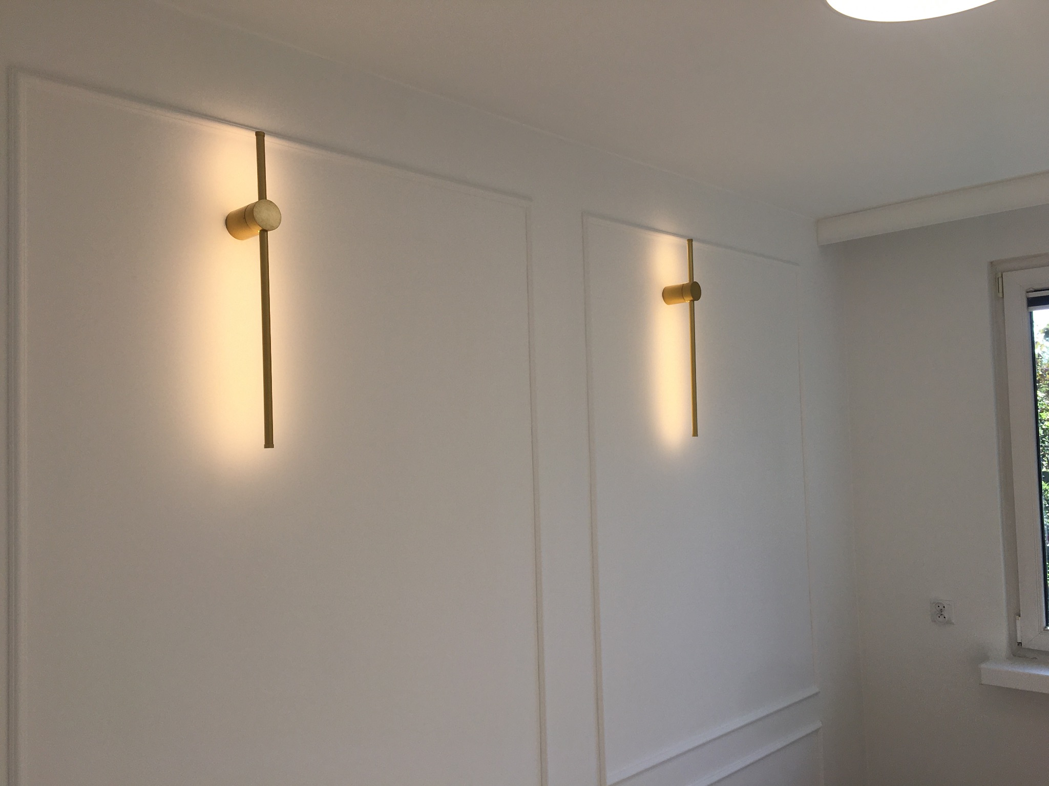 Białe ściany z ozdobnymi listwami i dwoma złotymi, podłużnymi kinkietami. W tle widoczne okno i fragment sufitu z oświetleniem.