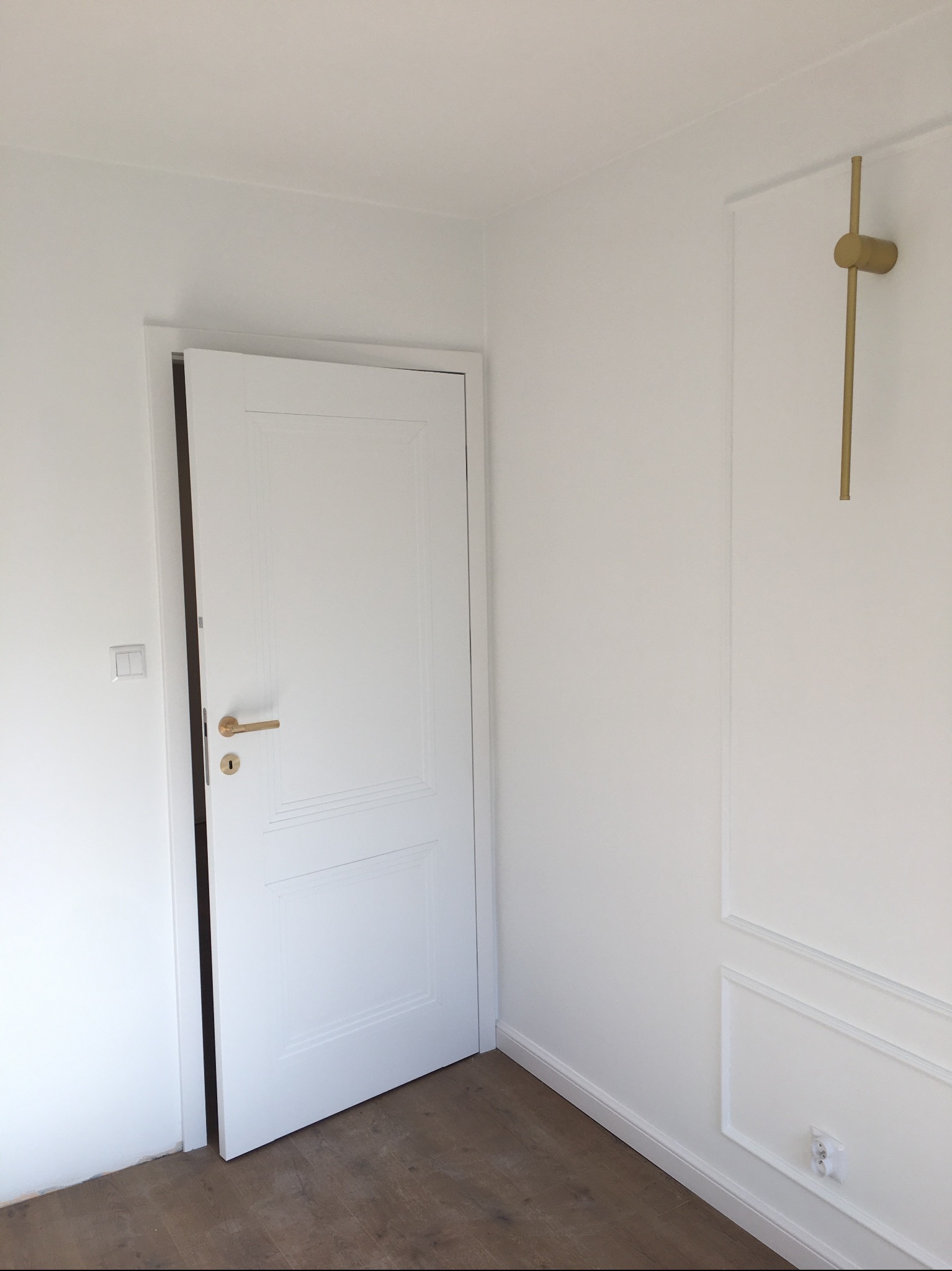 Białe, częściowo otwarte drzwi z złotą klamką w jasnym pokoju z dekoracyjną sztukaterią i drewnianą podłogą. Widoczny włącznik światła i gniazdko.