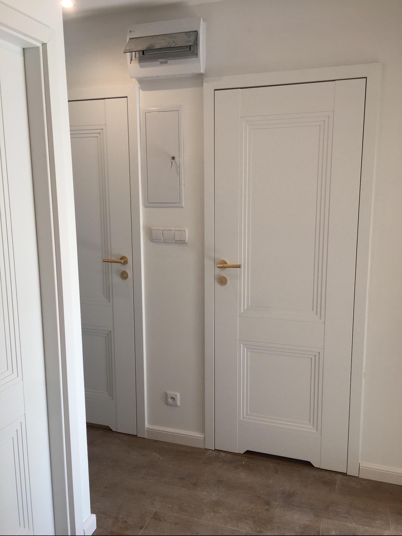 Dwa białe, panelowe drzwi z złotymi klamkami w jasnym wnętrzu. Jedne drzwi są uchylone, ukazując fragment podłogi i ściany. Nad drzwiami widoczna skrzynka elektryczna.