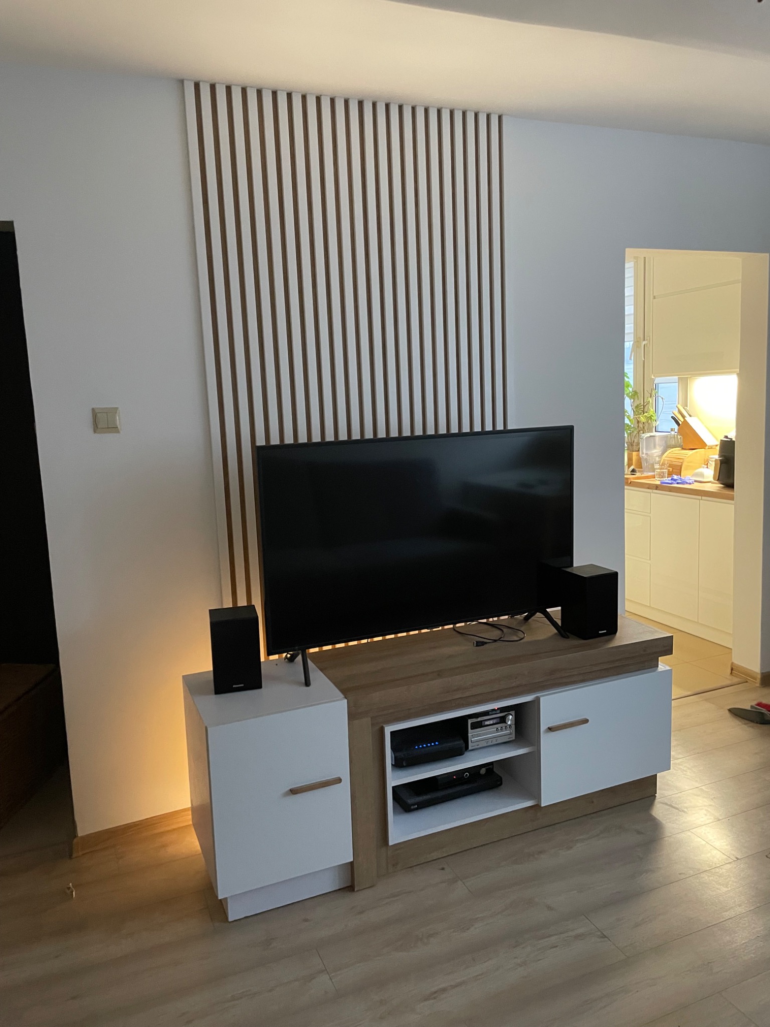 Nowoczesny salon z telewizorem na szafce RTV, drewniane lamele na ścianie i widok na kuchnię w tle. Minimalistyczny design, ciepłe oświetlenie.