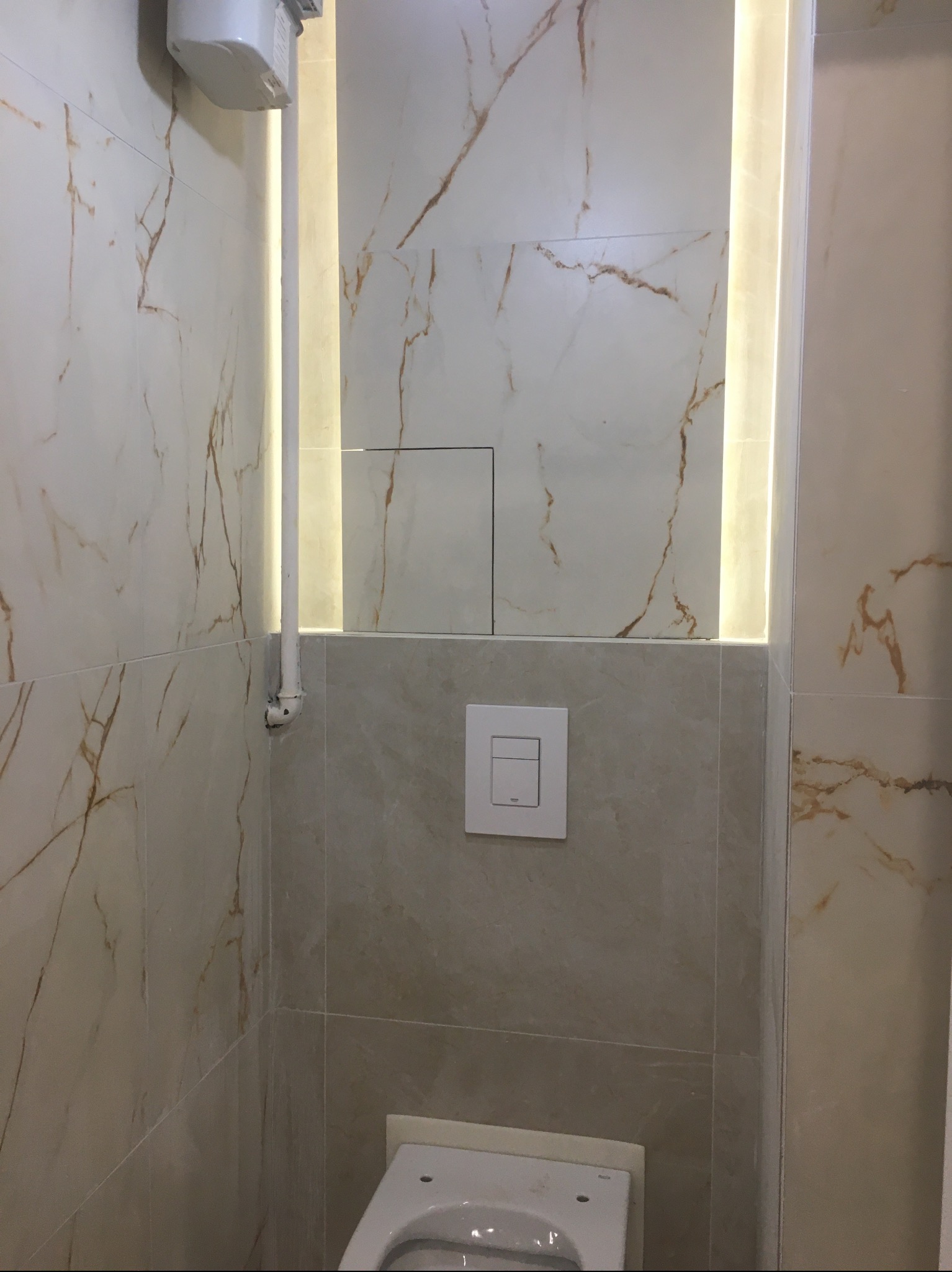 Wykończona łazienka z beżowymi płytkami imitującymi marmur, podświetleniem LED w pionowej wnęce i minimalistycznym panelem spłukującym. Widoczny fragment toalety.