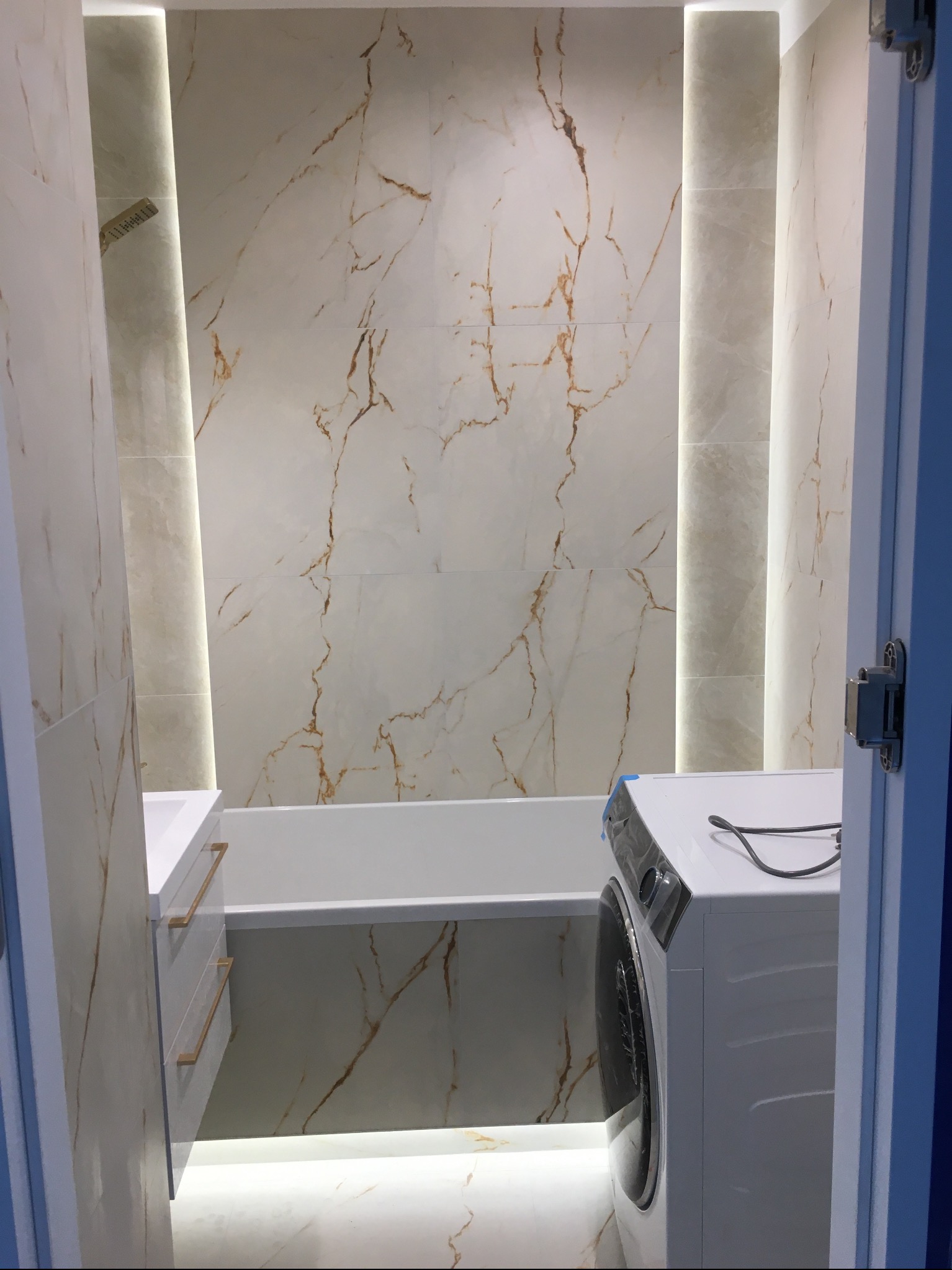 Wykończona łazienka z marmurowymi płytkami, podświetleniem LED, białą wanną i szafką pod umywalką. Widoczna pralka. Ujęcie zza uchylonych drzwi.