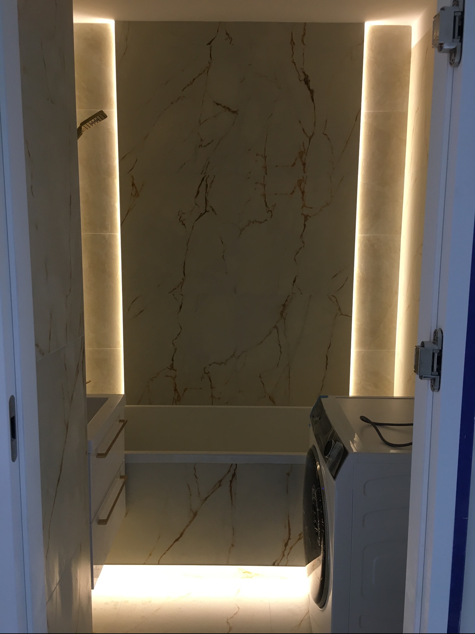 Nowoczesna łazienka z marmurowymi płytkami, podświetleniem LED i pralką. Widok zza uchylonych drzwi, ukazujący minimalistyczny design i dbałość o detale.