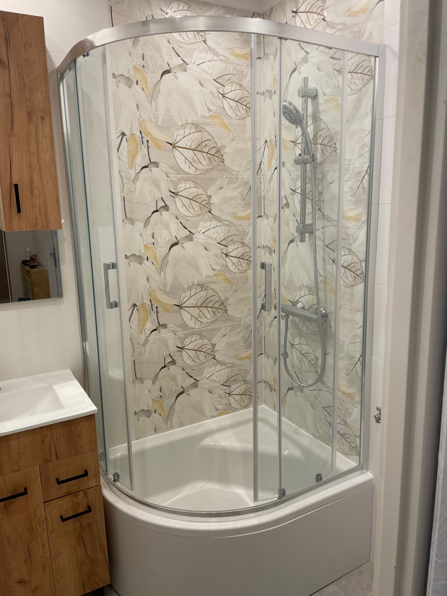 Wykończona łazienka: kabina prysznicowa z wzorem liści, drewniana szafka z umywalką. Nowoczesny design, jasne płytki, chromowane elementy.