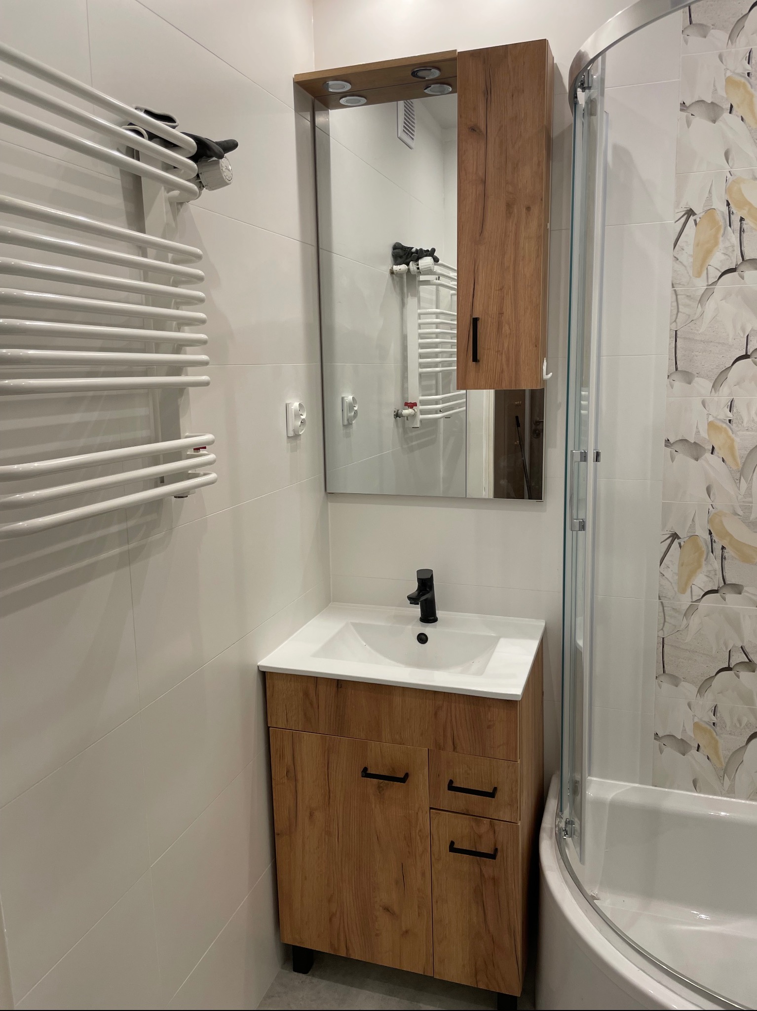 Nowoczesna łazienka po remoncie: białe płytki, szafka z umywalką w kolorze drewna, lustro z szafką i grzejnik drabinkowy. Kabina prysznicowa z widocznym wzorem na płytkach.