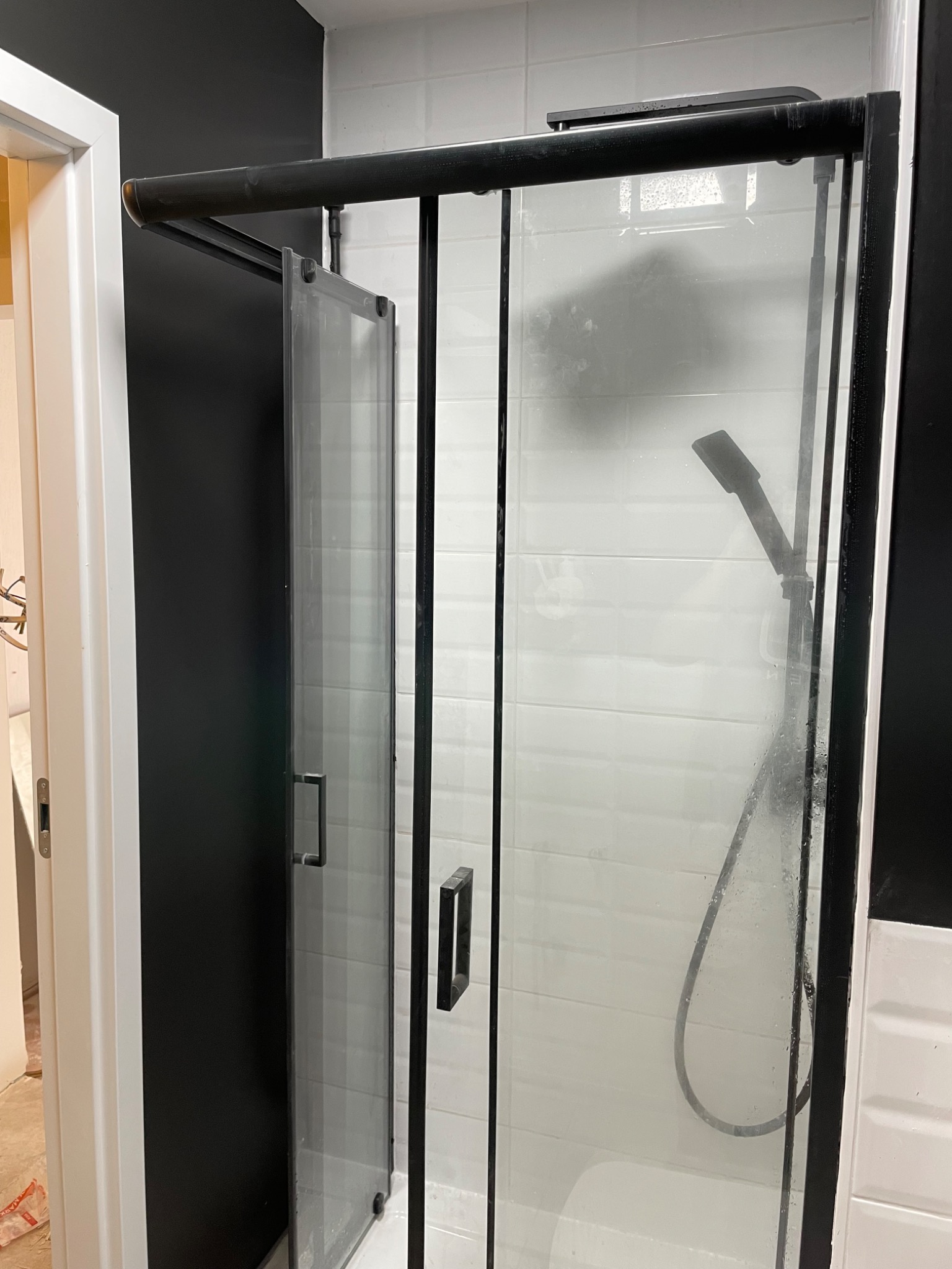 Nowoczesna kabina prysznicowa z czarnymi profilami i uchwytami, częściowo otwarta, na tle białych płytek i czarnej ściany. Widoczny fragment łazienki w trakcie wykończenia.