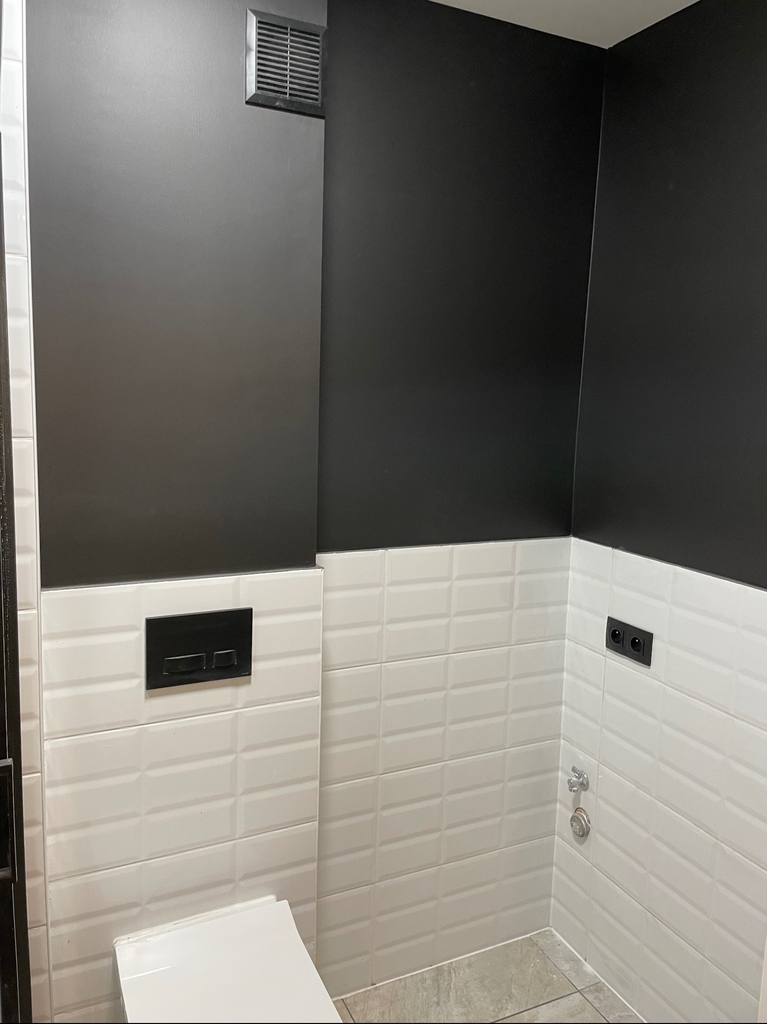 Nowoczesna łazienka po remoncie: białe, wypukłe płytki na ścianach kontrastują z czarną górną częścią ścian. Widoczny fragment toalety i czarny panel sterujący spłuczką.