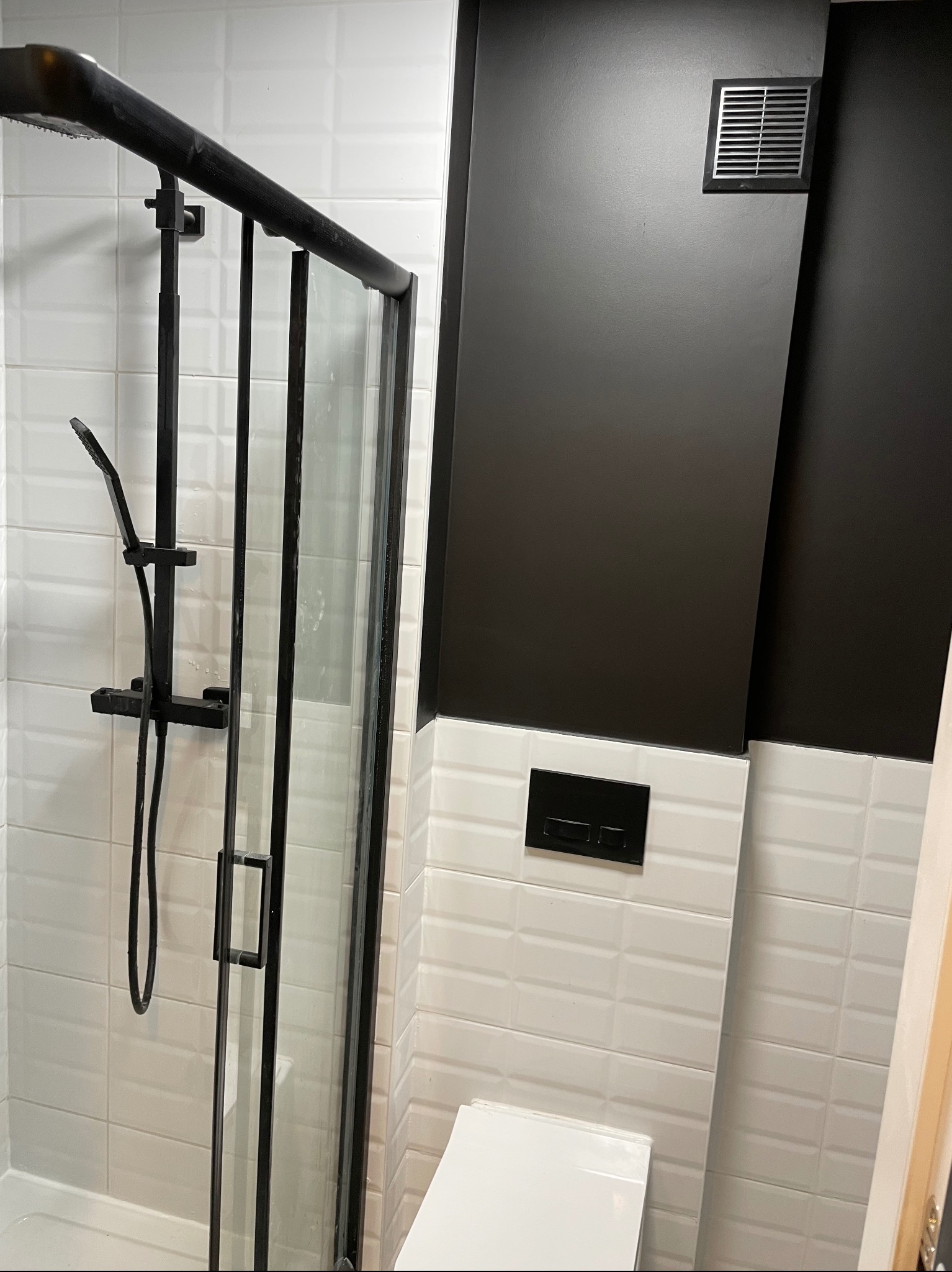 Nowoczesna łazienka z czarnymi akcentami: kabina prysznicowa typu walk-in, białe płytki imitujące cegłę i czarna ściana z wentylacją. Minimalistyczny design.