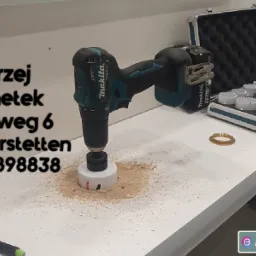 Wiertarka akumulatorowa Makita z otwornicą wierci otwór w białym blacie, widoczne wióry, zestaw otwornic w aluminiowej walizce i dane kontaktowe firmy na ścianie.