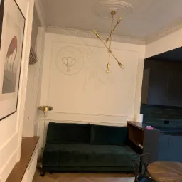 Elegancki salon z ciemnozieloną sofą, parkietem w jodełkę, nowoczesną lampą wiszącą i dekoracyjnymi sztukateriami na ścianach, w tle widoczna ciemna kuchnia.