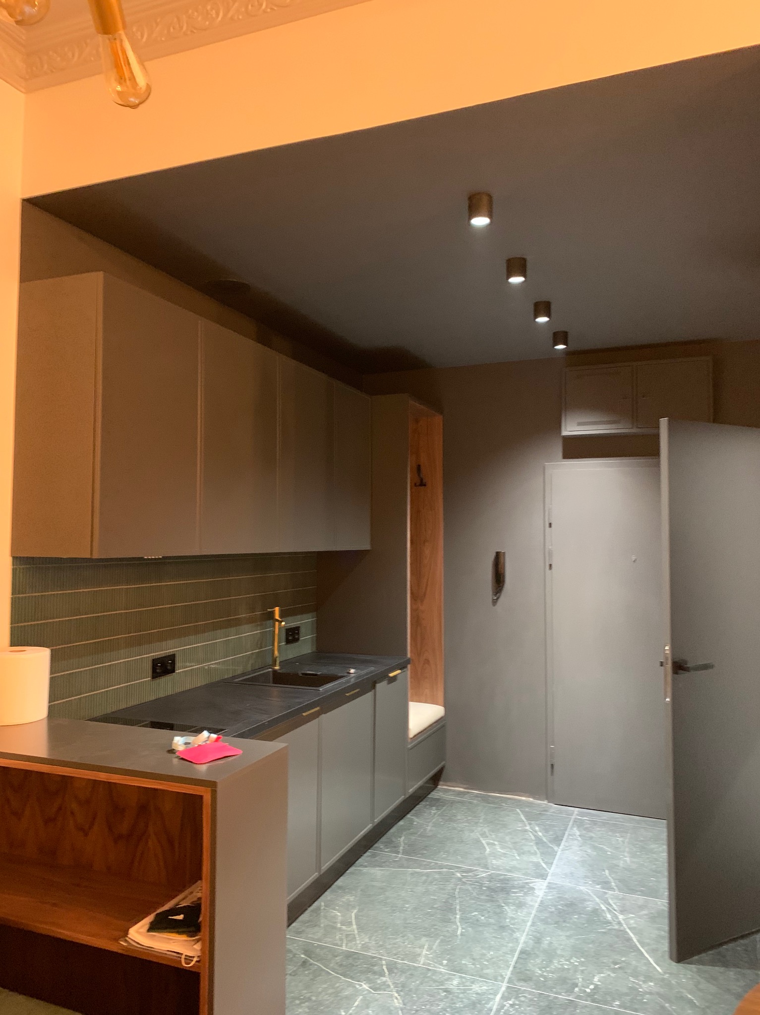 Nowoczesna kuchnia w odcieniach szarości z blatem z ciemnego kamienia, podłużnymi płytkami nad blatem i złotą baterią. Widoczne punktowe oświetlenie sufitowe i marmurowa podłoga. Częściowo otwarte...