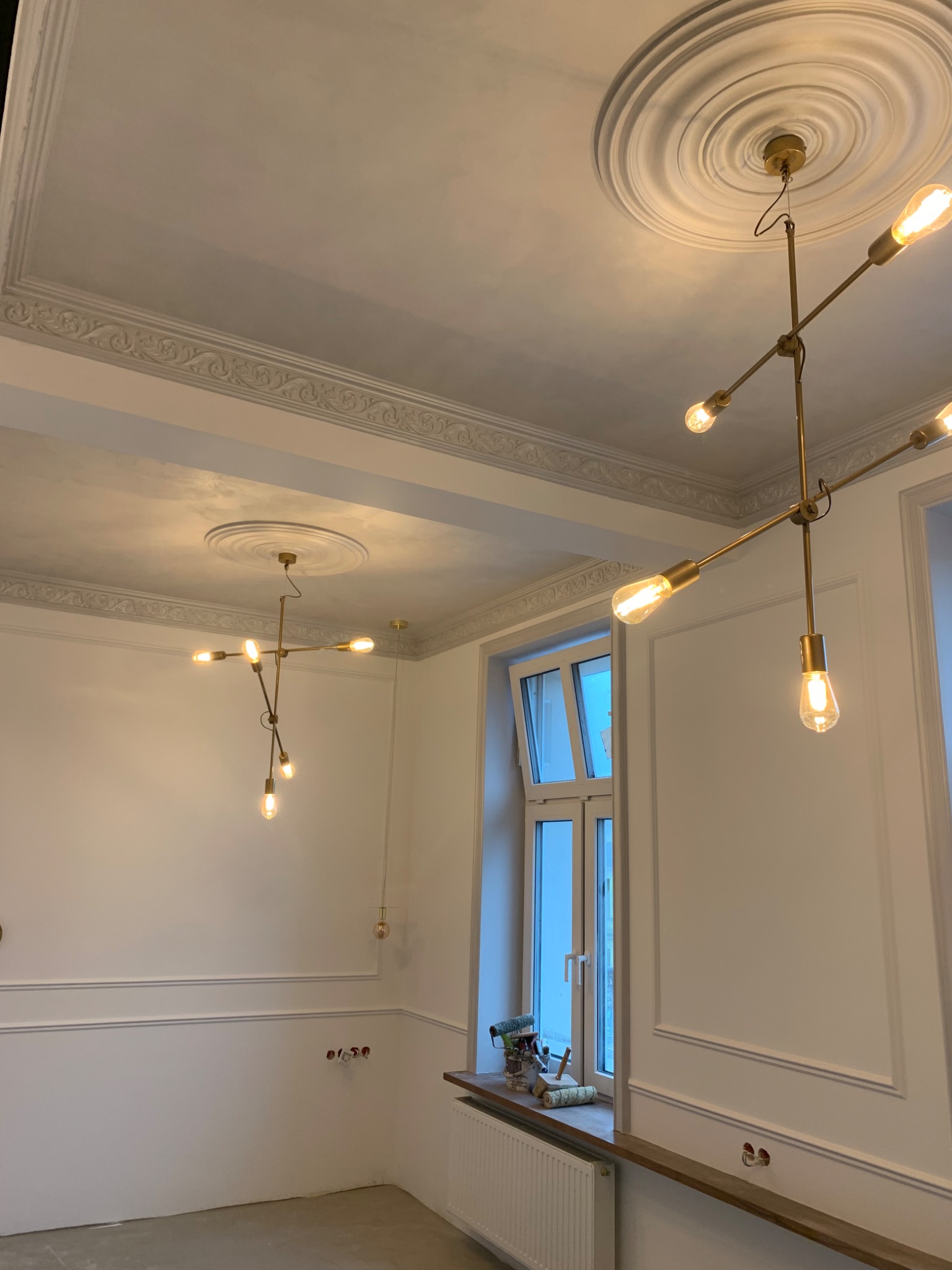Wnętrze w Szczecinie w trakcie prac wykończeniowych: widoczne listwy dekoracyjne na ścianach, ozdobny sufit z rozetą i nowoczesne, złote lampy z żarówkami Edisona, okno z narzędziami malarskimi...