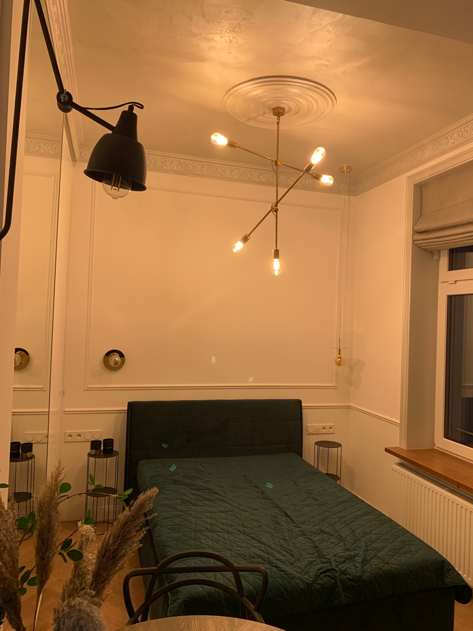 Sypialnia z zielonym łóżkiem, oświetlona lampą sufitową w stylu retro i czarną lampą ścienną, ściany z dekoracyjnymi listwami, widoczne elementy dekoracyjne i okno z żaluzjami.