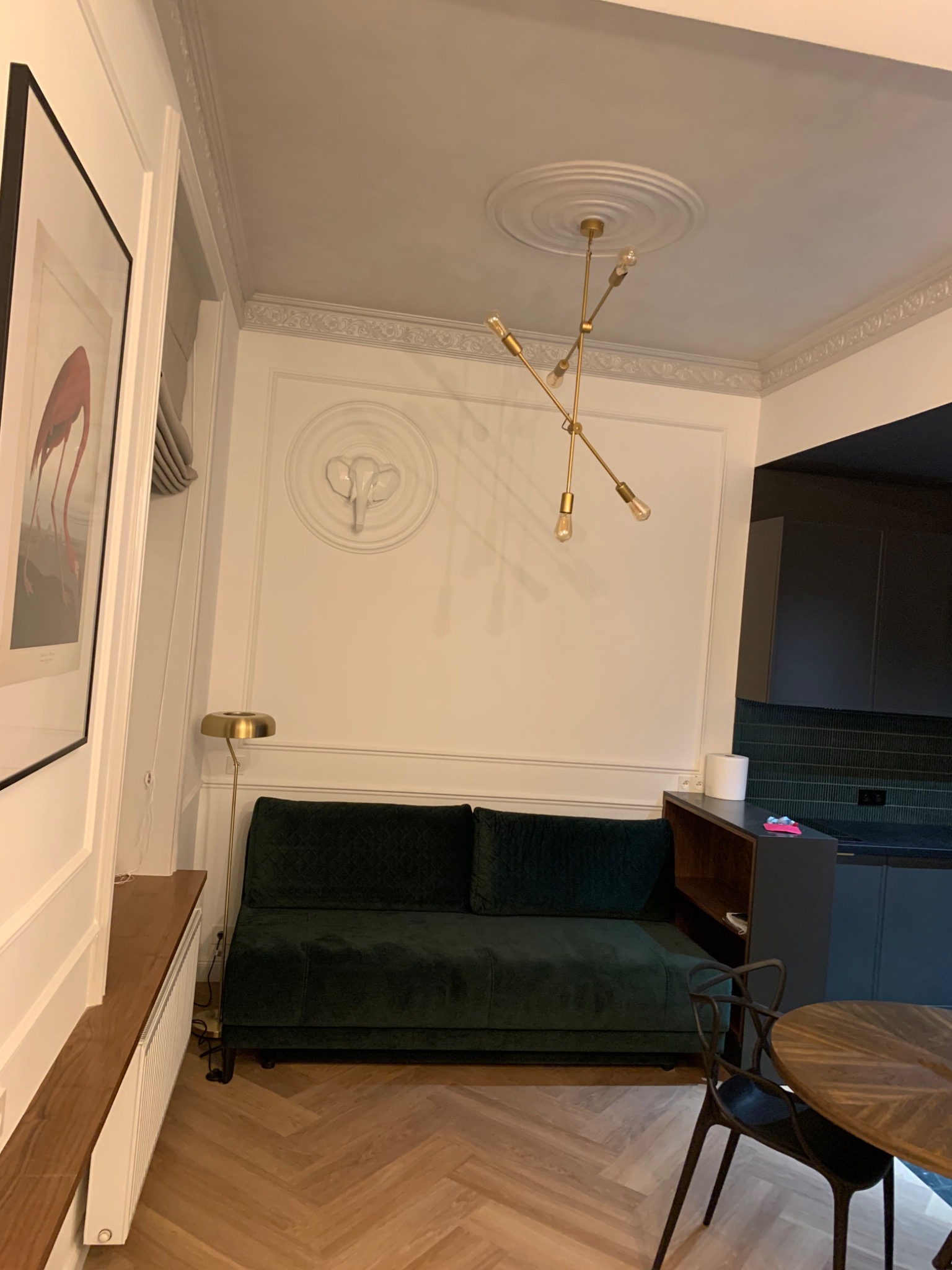 Elegancki salon z ciemnozieloną sofą, parkietem w jodełkę, nowoczesną lampą wiszącą i dekoracyjnymi sztukateriami na ścianach, w tle widoczna ciemna kuchnia.