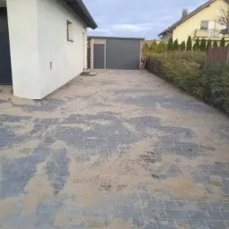 Nowo ułożona szara kostka brukowa z piaskiem na fugach, widok z perspektywy chodnika w kierunku garażu i domu z żółtą elewacją w tle.