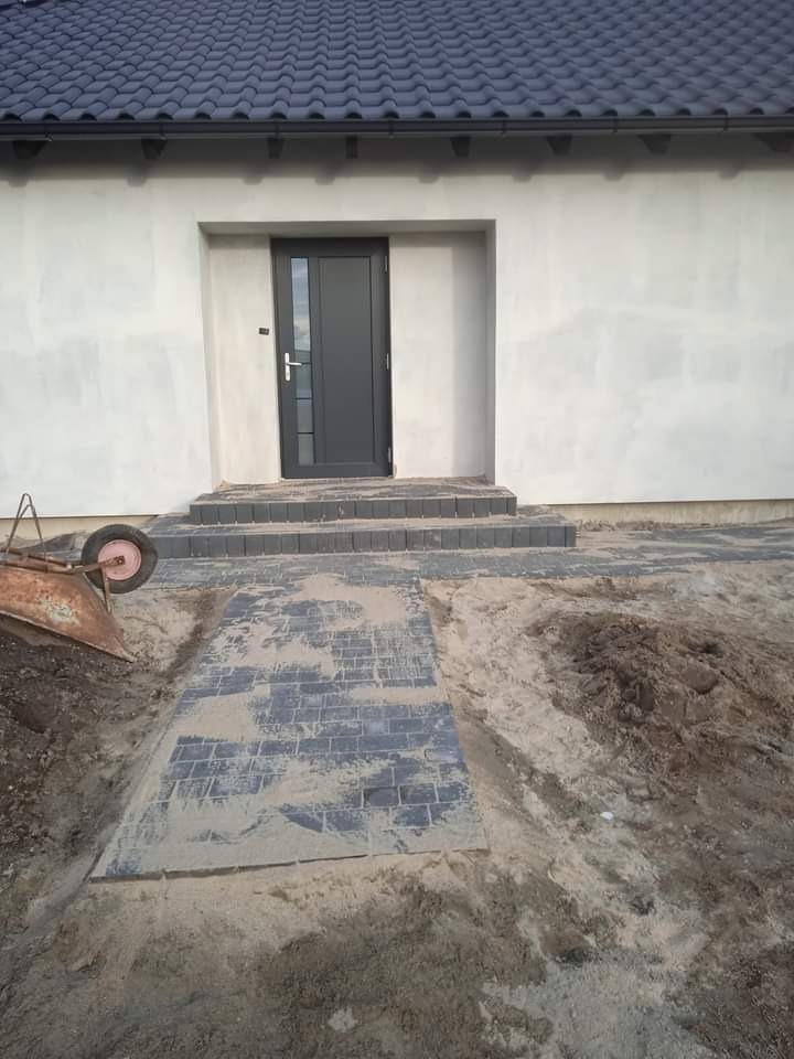 Ułożona kostka brukowa prowadząca do wejścia domu z szarą stolarką drzwiową; widoczny fragment taczki i piasek.