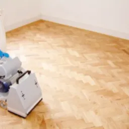 Parkiet ułożony we wzór jodełki, świeżo po cyklinowaniu, z widoczną maszyną do cyklinowania i workiem na pył w rogu pomieszczenia.