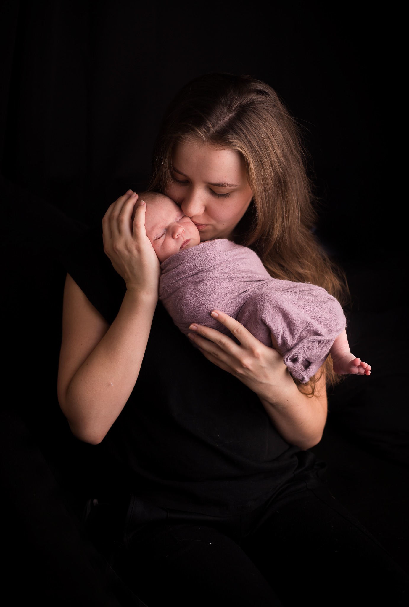 Matka całuje śpiące nowonarodzone dziecko owinięte w liliowy kocyk na tle ciemnego tła.